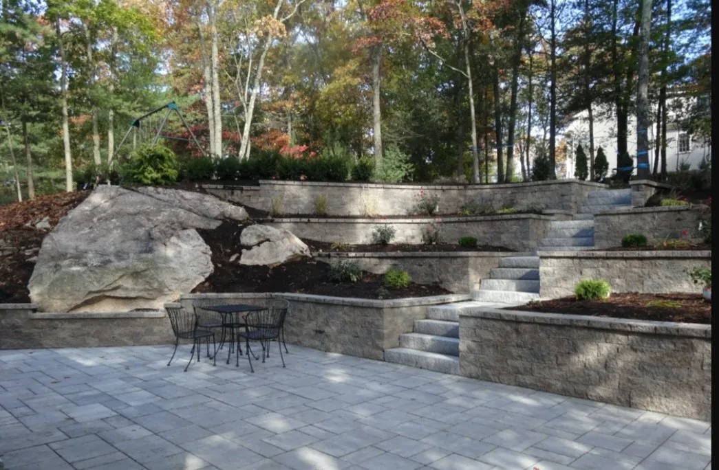 Terraced stone retaining walls with a paver patio and natural boulder in a landscaped backyard